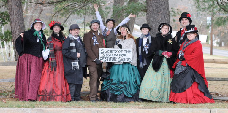 A photograph of a group of ten people dressed in Victorian garb posing together. A member near the center holds a sign that reads Society of the Asylum for Destitute Sailors