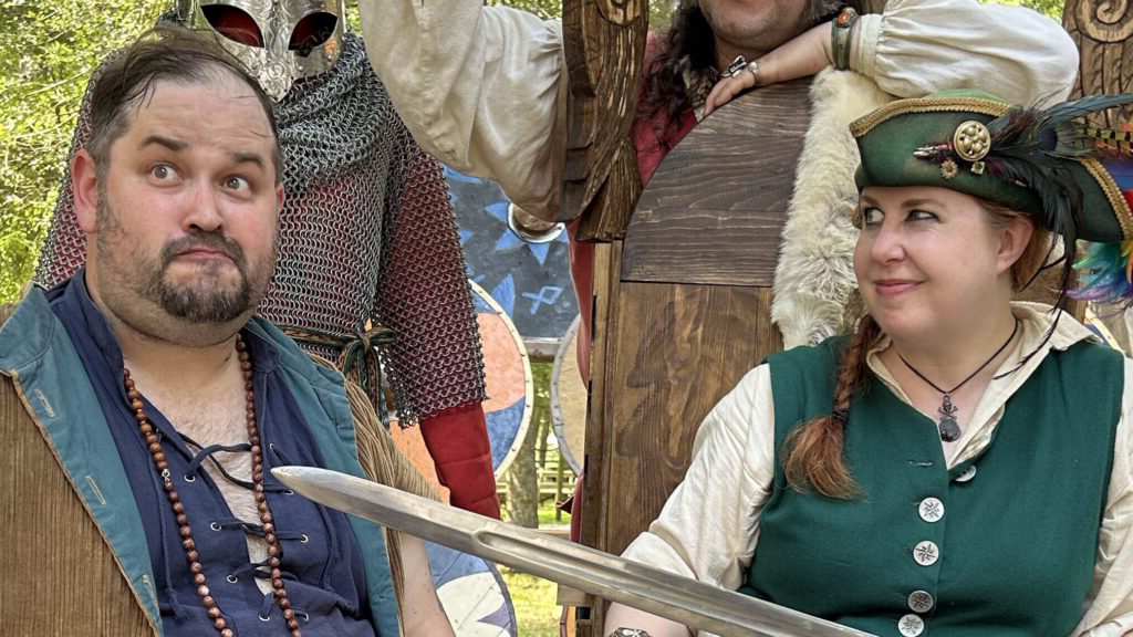 A photograph of a female dressed as a pirate smiling and playfully pointing a sword at the male dressed as a pirate beside her. He makes a silly expression and looks away from he and the sword