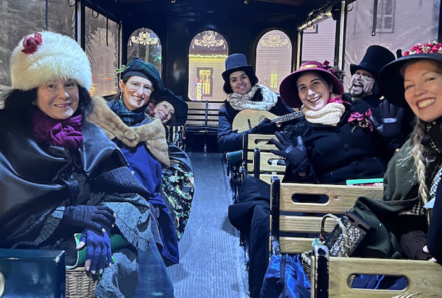 A photograph of a group of people dressed in Victorian garb smiling and sitting in the rows of a trolley