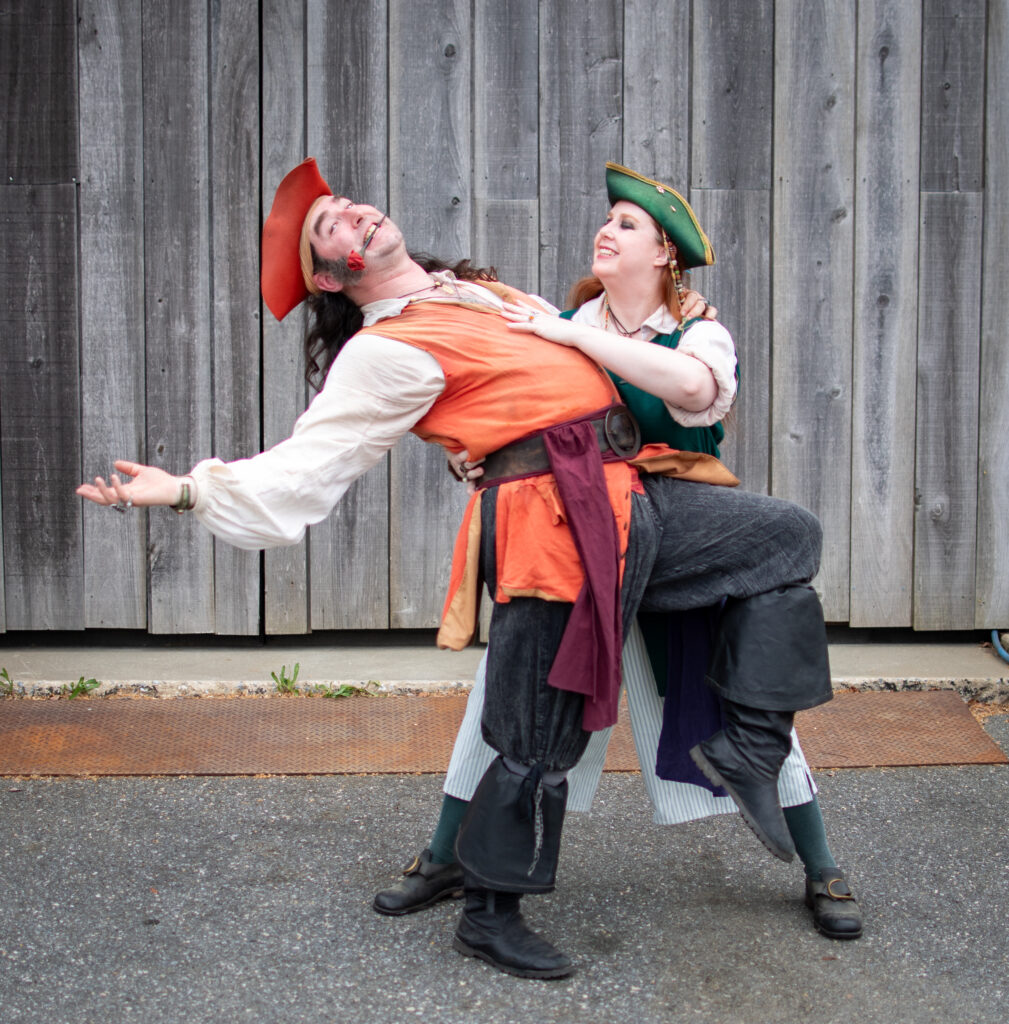 A photograph of a man dressed as a pirate and a female dressed as a pirate tangoing together. The female pirate dips the male down as he holds a rose in his mouth. They are both smiling