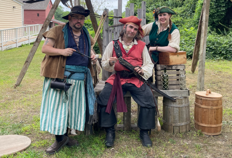A photograph of two people dressed as pirates sitting on a wooden throne-like chair, and a third pirate standing near the chair. All three people are posing in a tough manner