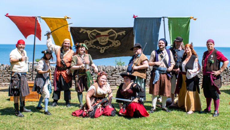 A photograph of eleven people dressed as pirates smiling and posing in front of a flag with the Pirates of Fortune's Folly logo, a skull with a jester hat and keys behind it instead of bones. Nine of the pirates are posing with objects such as bladed weapons and eighteenth-century style mugs