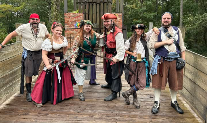 A photograph of six people dressed as pirates standing on a bridge. They are all smiling and four of them are posing with bladed weapons.