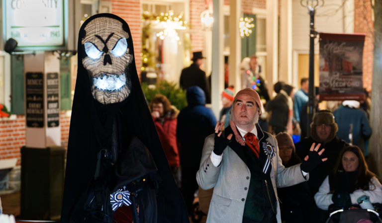 A photograph of a man dressed in Victorian garb looking afraid of a large glowing skull representing the Ghost of Christmas Yet to Come