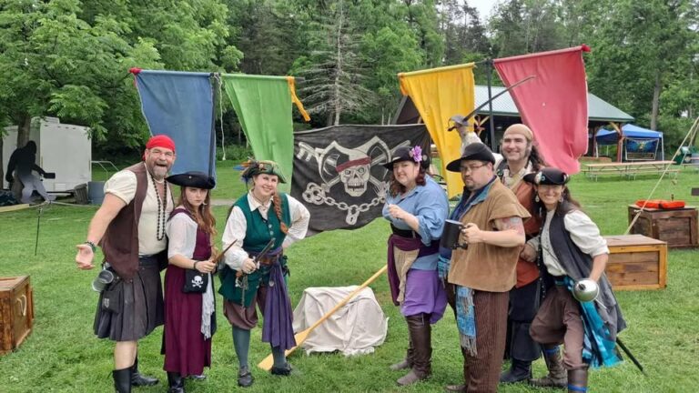 A photograph of seven people dressed as pirates posing in front of a flag with the Pirates of Fortune's Folly logo, a skull with a jester hat and keys behind it instead of bones. Four of the pirates pose with bladed weapons, one poses with a comically large fork, and one has an eighteenth-century mug