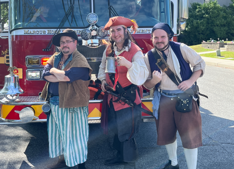 A photograph of three men dressed as pirates smiling and standing in front of a fire truck