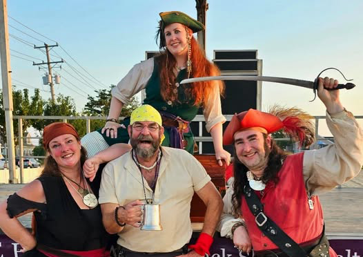 A photograph of two men and two women dressed as pirates posing together and smiling. One man holds an eighteenth-century style mug and the other man holds a sword above his head