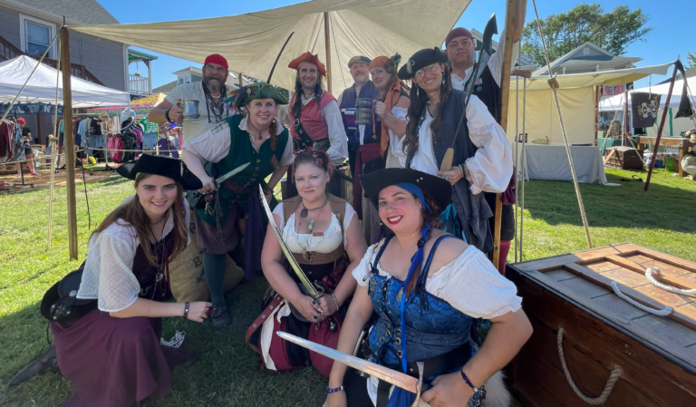 A photograph of ten people dressed as pirates smiling and standing under the shade of a canopy. Four of the pirates in the foreground and midground hold bladed weapons