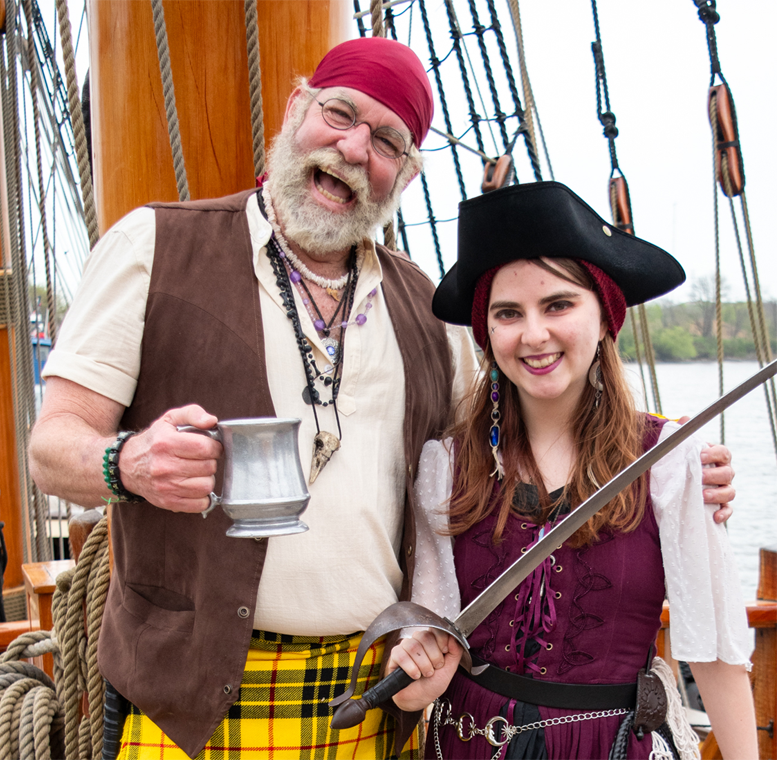 A photograph of a man dressed as a pirate holding an eighteenth-century style mug in one hand and putting his other hand around a female dressed as a pirate. She is holding a sword. They are both standing on a pirate ship