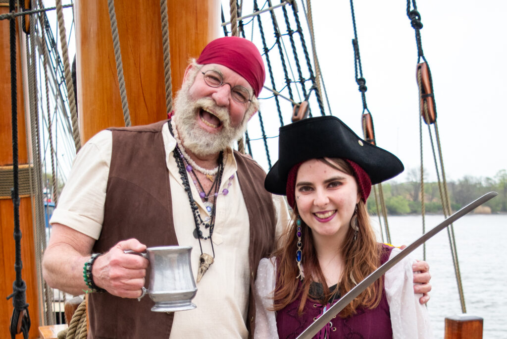 A photograph of a man dressed as a pirate holding an eighteenth-century style mug in one hand and putting his other hand around a female dressed as a pirate. She is holding a sword. They are both standing on a pirate ship