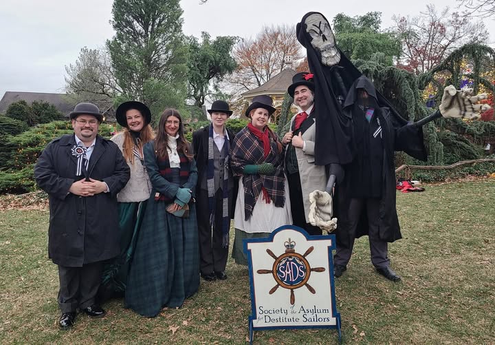 A photograph of a group of people standing in a grassy area behind a sign that reads Society of the Asylum for Destitute Sailors. Six people are dressed in Victorian garb, one is dressed in a large costume with a skull representing the Ghost of Christmas Yet to Come