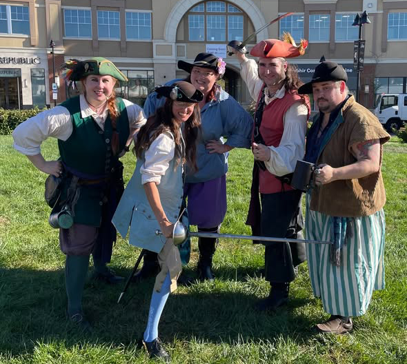 A photograph of five people dressed as pirates standing on grass in front of a mall, two of them are posing with swords and one has an eighteenth-century style mug