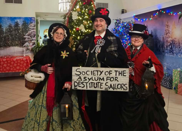 A photograph of three people dressed in Victorian garb standing in front of a Christmas tree smiling. The person in the center holds a sign that reads Society of the Asylum for Destitute Sailors